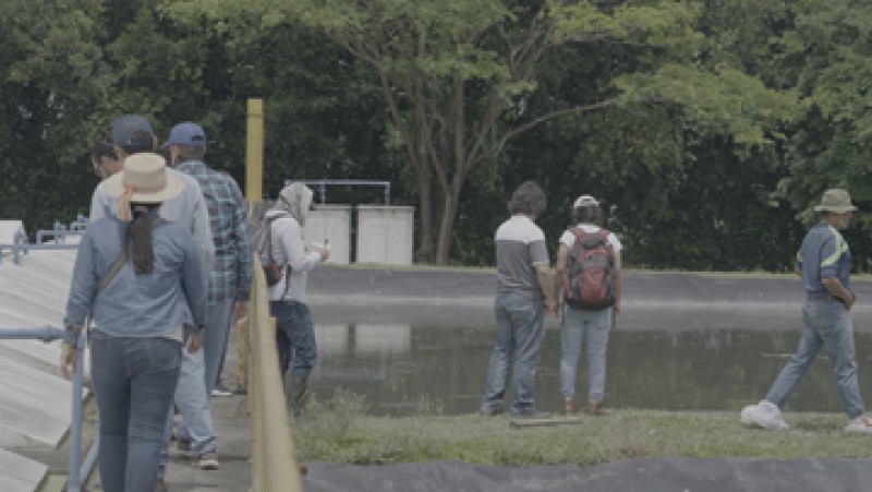 Course participants walk alongside water tanks and equipment, and a riverbank where dark green trees overhang the water